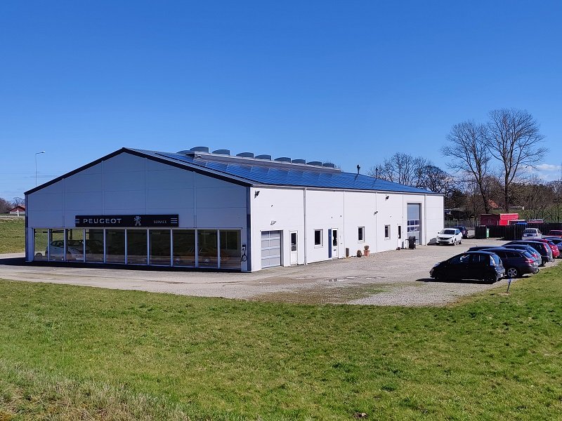 en lagerhal med solcellepaneler på taget omringet af græsmark og en parkeringsplads med biler