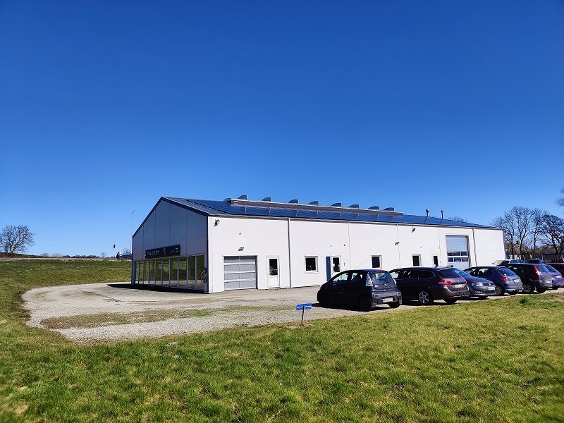 en lagerhal med solcellepaneler på taget omringet af græsmark og en parkeringsplads med biler
